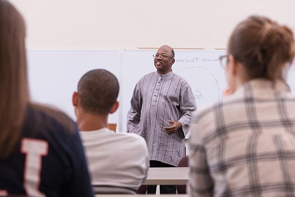 A smiling male professor teaching a group of students in front of a whiteboard during a religious studies class